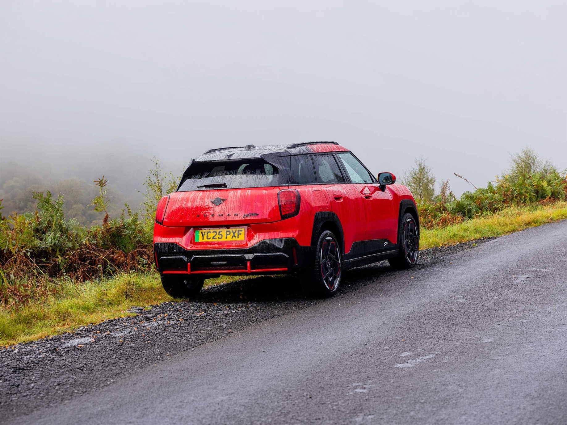Rear side view of parked all-electric MINI Aceman in British Racing Green in car park with all doors open and coloured wall in the background